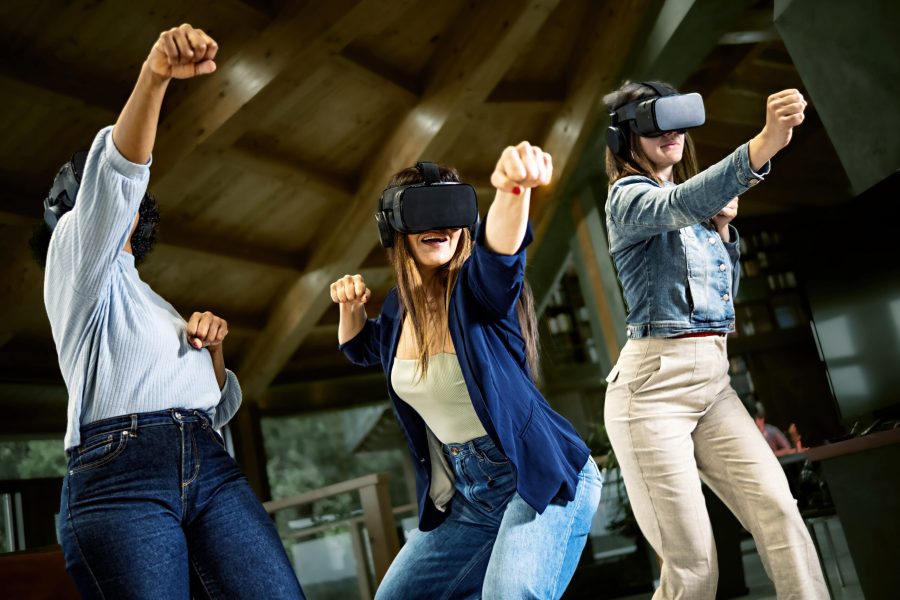Group of young women using virtual reality headset - Girlfriends playing ninja fight video games in meta verse space with futuristic goggles having fun together